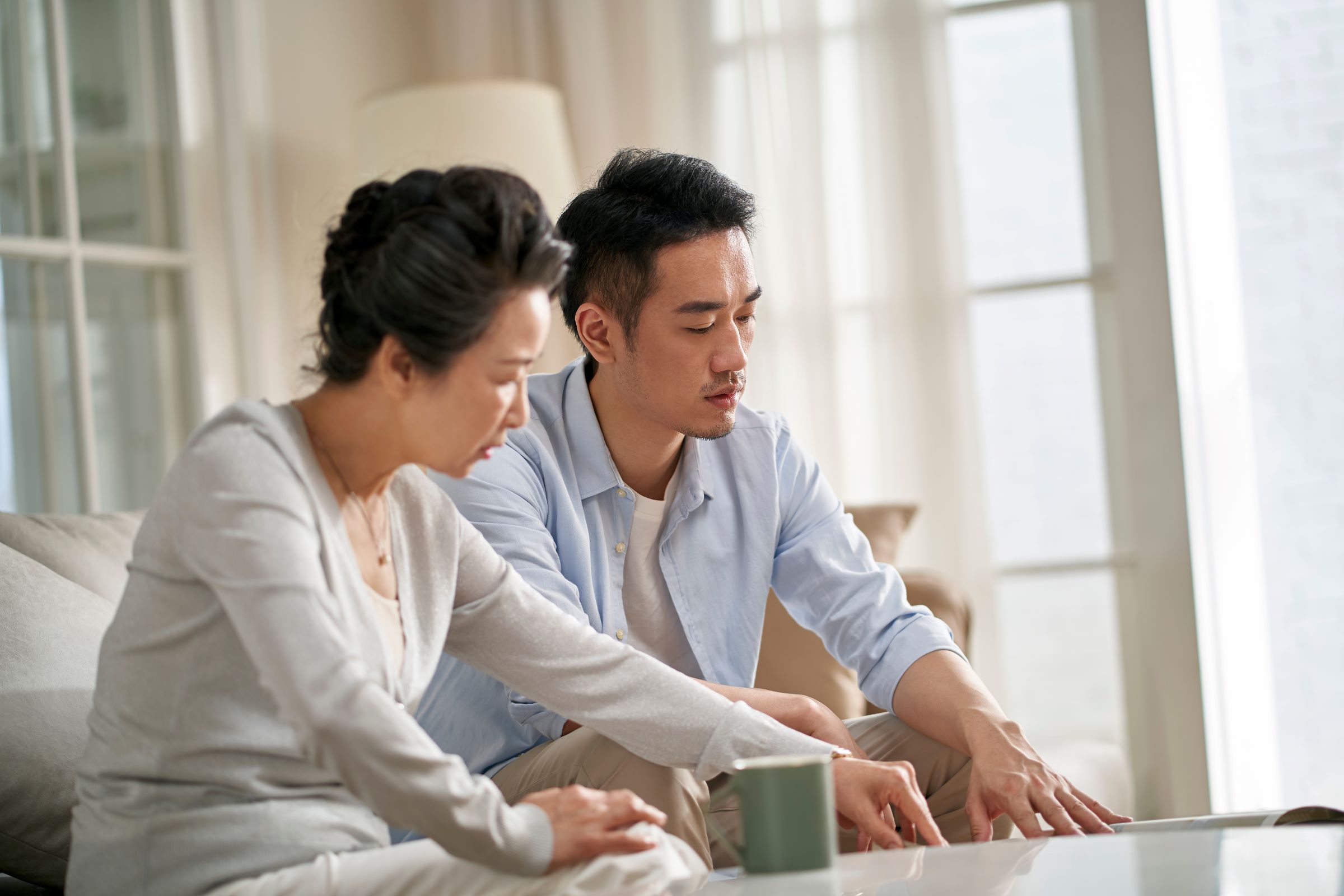 Senior mother sits on couch with her adult son having thoughtful conversation about finances.